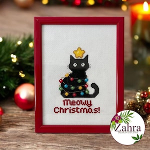 Könnte beinhalten: Eine Kreuzstichstickerei in einem roten Rahmen, die eine schwarze Katze zeigt, welche in bunte Lichterketten eingewickelt ist und einen gelben Stern auf dem Kopf trägt. Darunter steht der Text Meowy Christmas! in roten Buchstaben auf weißem Stoff.