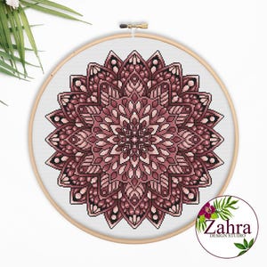 May include: A circular wooden embroidery hoop containing a cross-stitch pattern of a symmetrical mandala in shades of dusty rose, mauve, and dark burgundy on a white fabric background. A circular logo with the text Zahra Design Studio is visible.