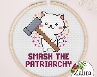 May include: A cross-stitch embroidery in a round wooden hoop featuring a white cat holding a gray mallet. The design includes pink and orange star-shaped accents and the text SMASH THE PATRIARCHY in dark purple block letters on white fabric.
