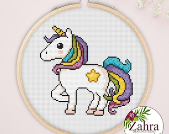 Puede incluir: Un proyecto de bordado en punto de cruz que muestra un unicornio blanco con crin y cola multicolor, una estrella amarilla en el flanco y un cuerno dorado, presentado en un bastidor de madera redondo sobre un fondo claro.