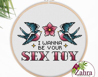 Könnte beinhalten: Ein runder Stickrahmen aus Holz mit einem Kreuzstichmotiv auf weißem Stoff. Das Design zeigt zwei blaue und rosafarbene Schwalben neben einer rosa Blume sowie den schwarzen Text I WANNA BE YOUR und den großen rosa Text SEX TOY.