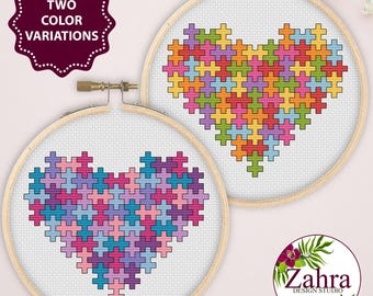 May include: Two circular wooden embroidery hoops containing cross-stitch patterns of hearts made from colorful puzzle pieces. One heart features a multi-colored palette, while the other uses shades of pink, purple, and blue. Text reads TWO COLOR VARIATIONS.