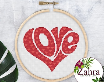 May include: A circular wooden embroidery hoop containing white fabric with a cross-stitch design of the word LOVE shaped into a heart. The letters are filled with a repeating pattern of small red hearts on a darker red background.