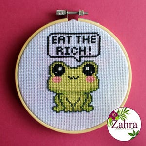 Op de afbeelding: Een borduurring met een wit achtergrond en een groene kikker met grote ogen. De kikker houdt een spreekballon vast met de tekst "Eat the Rich!"