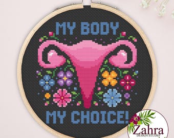 May include: A circular wooden embroidery hoop containing a cross-stitch design on black fabric. The design features a pink uterus illustration surrounded by colorful flowers and the blue text MY BODY MY CHOICE! in a blocky, pixelated font style.