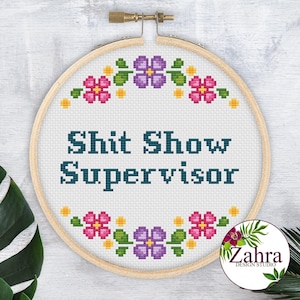 May include: A circular wooden embroidery hoop containing white fabric with cross-stitched text that reads "Shit Show Supervisor" in dark teal. The text is framed by a floral pattern of purple and pink flowers with green leaves and yellow accents.