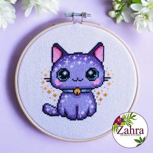 Puede incluir: Un bordado de punto de cruz en un bastidor de madera circular que muestra un gato morado con grandes ojos azules, orejas rosadas y un collar azul con un cascabel dorado. El gato tiene un patrón galáctico con estrellas blancas y doradas.