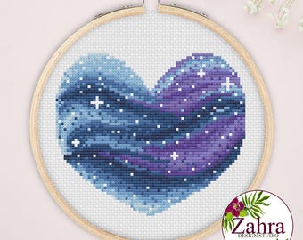 May include: A cross-stitch pattern of a heart shape featuring a galaxy design with shades of blue and purple, accented by small white stars. The embroidery is held within a round wooden hoop with a metal tightening screw at the top.