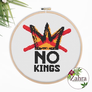 Op de afbeelding: Een borduurring met kruissteek met een kroonontwerp in rood, geel en zwart, met de woorden "NO KINGS" in zwarte blokletters. De borduursel staat tegen een witte achtergrond in een houten ring.