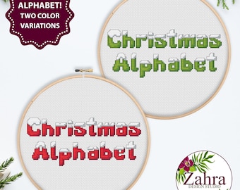 May include: Two circular wooden embroidery hoops containing cross-stitch patterns on white fabric. Each hoop displays the text "Christmas Alphabet" in a pixelated font with white snow-like tops, one in green and one in red color variations.