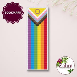 May include: A rectangular cross-stitch bookmark featuring a vertical rainbow stripe pattern. At the top, a triangular chevron design incorporates brown, black, light blue, pink, and white colors, with a small yellow circle centered within the triangle.
