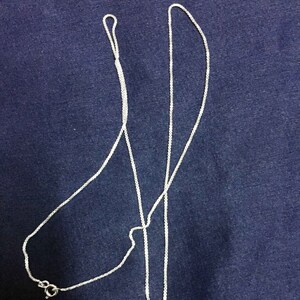May include: A silver chain necklace with a delicate, box chain design. The chain is laid out on a blue fabric background.