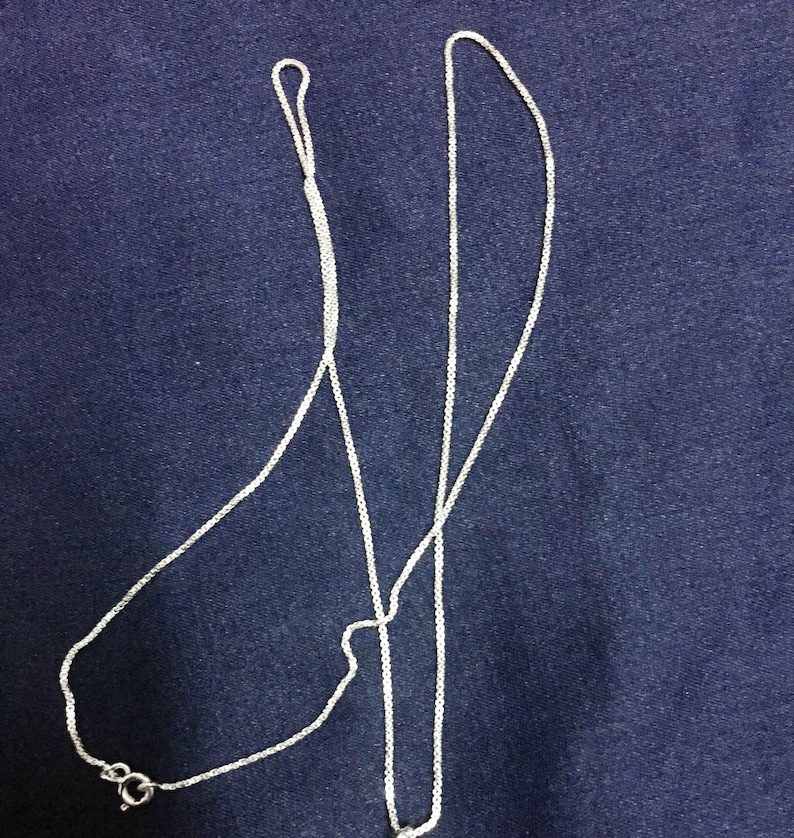 May include: A silver chain necklace with a delicate, box chain design. The chain is laid out on a blue fabric background.