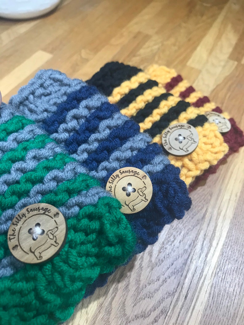 May include: Four knitted cup cozies in different color combinations, each with a wooden button featuring a dachshund and the text "The Silly Sausage".