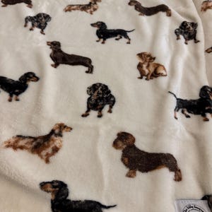 May include: A cream-colored blanket featuring a repeating pattern of dachshund dogs in various poses and colors, including black, brown, and tan. The blanket has a soft, plush texture and a small tag with a logo in the corner.