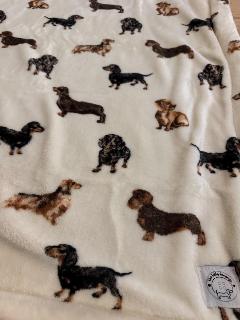 May include: A cream-colored blanket featuring a repeating pattern of dachshund dogs in various poses and colors, including black, brown, and tan. The blanket has a soft, plush texture and a small tag with a logo in the corner.