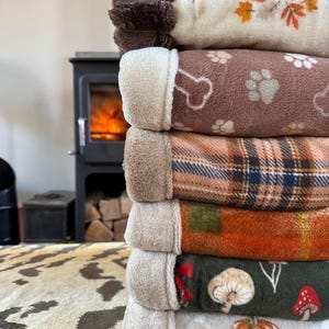 May include: A vertical stack of six folded plush blankets. The top blanket is cream with orange leaves, followed by a brown blanket with white paw prints and bones, a plaid blanket, an orange plaid blanket, a dark green blanket with mushrooms, and a cream blanket with orange pumpkins.