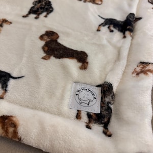 May include: Cream-colored plush blanket featuring a repeating pattern of dachshund dogs in various colors. The blanket has a soft, textured appearance and a small label with the text "The Silly Sausage" and a dachshund illustration.