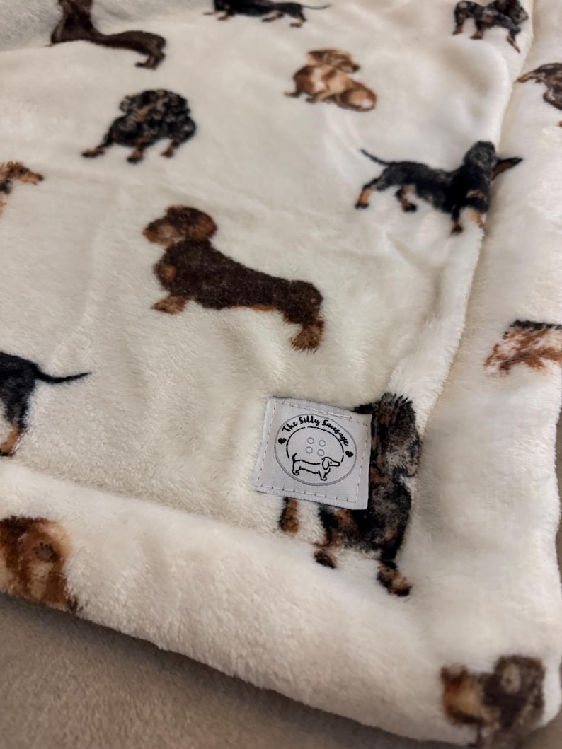 May include: Cream-colored plush blanket featuring a repeating pattern of dachshund dogs in various colors. The blanket has a soft, textured appearance and a small label with the text "The Silly Sausage" and a dachshund illustration.