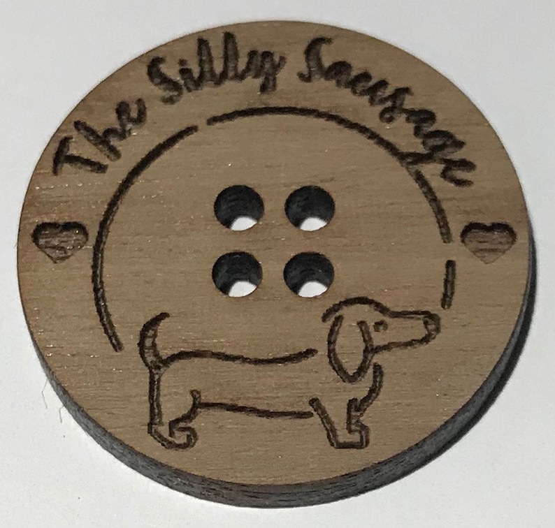 May include: A wooden button with four holes and a dachshund silhouette. The button is engraved with the text "The Silly Sausage".