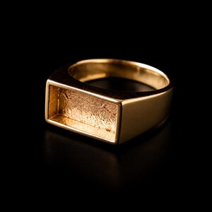 Könnte beinhalten: Ein goldfarbener Ring mit einer rechteckigen Vorderseite und einer vertieften, strukturierten Mitte. Das Band ist glatt und poliert und weist ein klares, geometrisches Design auf. Die Fassung ist für eine Einlage vorgesehen.