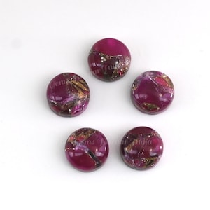 May include: Five round, polished gemstones with a deep magenta hue, interspersed with gold and silver flecks. The gemstones are arranged on a white surface. The text "Gems Bazaar India" is visible on some of the gemstones.