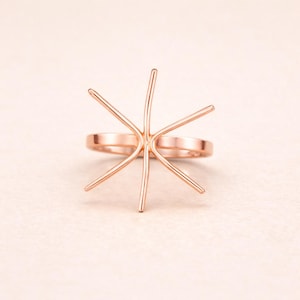 May include: A rose gold ring with a distinctive starburst design. The ring has a band and several prongs radiating outwards, forming a star-like shape. The ring is set against a soft, light pink background.