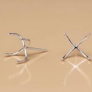 May include: Two silver-colored jewelry settings, one with three curved prongs and a post, the other with four crossed prongs. The settings are displayed on a light brown surface, reflecting their shapes.