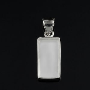Puede incluir: Un colgante rectangular con un centro blanco opaco y un marco de metal plateado. El colgante tiene un pequeño lazo en la parte superior para una cadena. El fondo es negro.