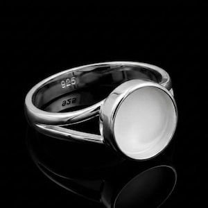 May include: A silver ring with a large, round, white stone. The ring has a split band design and is marked with "925" and "asa". The ring is set against a black background.