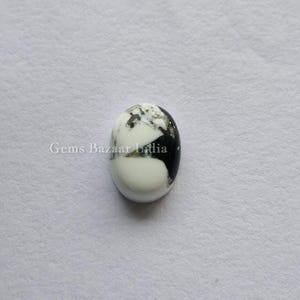 Puede incluir: Gema ovalada con base blanca, con vetas negras y grises. La piedra pulida tiene una superficie lisa y está sobre un fondo blanco. La gema es una piedra natural.