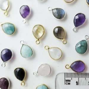 Può includere: Assortimento di ciondoli a goccia di pietra preziosa con accenti argentati e dorati. I ciondoli sono disposti su una superficie bianca con un righello per la scala. I ciondoli misurano circa 1 cm.