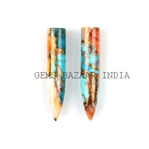 Może przedstawiać: Dwa turkusowe, pomarańczowe i złote szpiczaste kamienie szlachetne. Szpiczaste kamienie mają około 5 cm długości i zakończone są ostrym końcem.  Gems Bazaar India jest nadrukowane na białym tle.