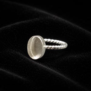Può includere: Un anello d'argento con una fascia intrecciata e un castone ovale. L'anello è realizzato in metallo lucido ed è progettato per contenere una pietra o un altro ornamento. L'anello è mostrato su uno sfondo nero.