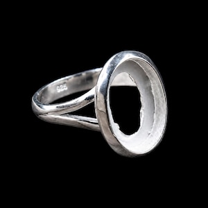 May include: A silver ring with an oval setting, perfect for a custom gemstone. The ring has a split shank design and is made of polished metal. The ring is shown against a black background.