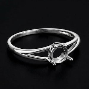 May include: A silver ring with a split shank design and a round setting. The ring is made of a shiny, polished metal and is set against a black background. The setting is designed to hold a gemstone.