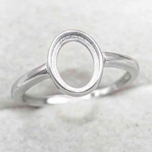 Puede incluir: Un anillo plateado con un engaste ovalado. El anillo tiene una banda sencilla y un bisel elevado para sujetar una piedra u otro adorno. El anillo se muestra sobre una superficie de color claro.