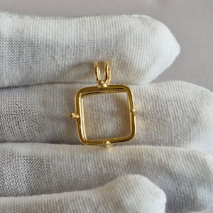 May include: A gold-colored pendant with a square frame and a loop at the top for a chain. The pendant has small decorative details at each corner. The background is a white fabric.