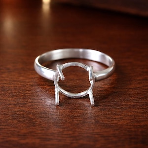 Peut inclure: Une monture de bague en argent avec un espace circulaire pour une pierre précieuse. La bague a une finition polie et une bande simple. Le sertissage comporte des griffes pour maintenir une pierre en place. La bague est présentée sur une surface en bois brun foncé.