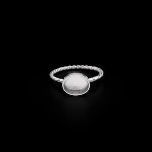 Könnte beinhalten: Ein silberner Ring mit einem gedrehten Band und einer kreisförmigen Lünettenfassung. Der Ring ist vor einem schwarzen Hintergrund platziert, der seinen metallischen Glanz hervorhebt. Die Lünette ist bereit, einen Stein oder ein anderes dekoratives Element zu halten.