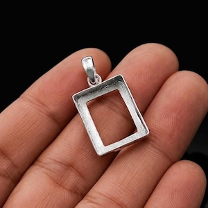Peut inclure: Un pendentif rectangulaire argenté avec une boucle en haut. Le pendentif a un centre creux, créant un design en forme de cadre. Le pendentif est tenu dans une main sur un fond noir.