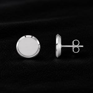 May include: A pair of silver-toned stud earrings featuring a circular, flat-faced design with a smooth, metallic finish. One earring is shown from the front, while the other is displayed from the side, revealing the post and butterfly clutch backing.