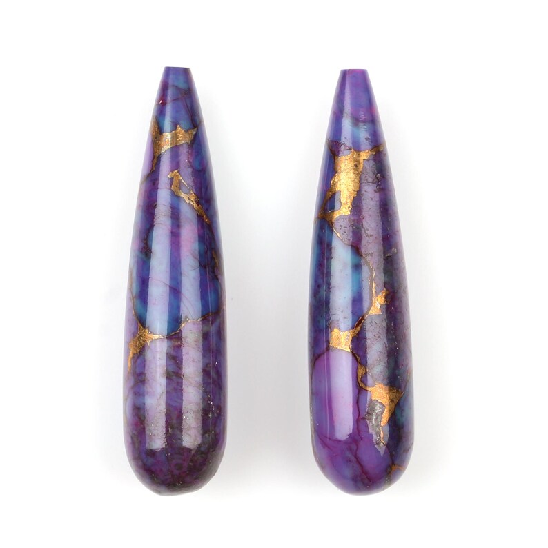 May include: Two teardrop-shaped purple and gold gemstone beads. The beads have a marbled pattern with gold flecks throughout.