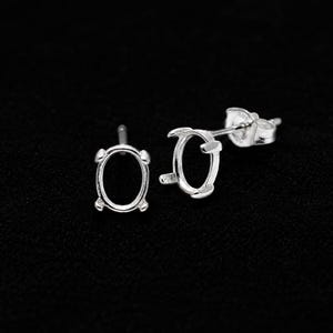 May include: A pair of silver stud earrings featuring oval-shaped prong settings designed to hold a gemstone. Each earring has a post back and a polished metallic finish, showcasing a simple frame structure intended for mounting a single oval stone.