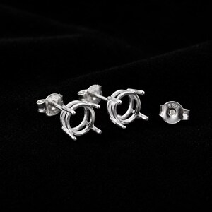 May include: A pair of sterling silver earring settings featuring a round prong mount design for gemstones. Each stud includes a post and a butterfly back stamped with 925, indicating the silver content. The metal has a polished, reflective finish.