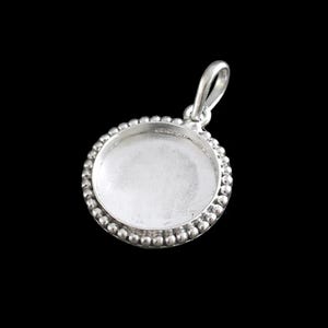 May include: A silver pendant with a circular, recessed centre and a beaded border. It has a loop at the top for a chain. The background is black.