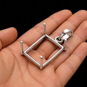 May include: A silver-colored pendant setting with a square frame and four vertical prongs. The setting includes a loop for a chain and is held in a hand against a black background.
