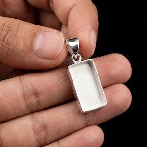 Könnte beinhalten: Rechteckiger Silberanhänger mit einer Schlaufe oben für eine Kette. Der Anhänger hat eine vertiefte Fläche, wahrscheinlich für einen Stein oder ein anderes dekoratives Element. Der Anhänger wird in einer Hand vor schwarzem Hintergrund gehalten.