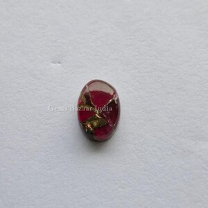 May include: Oval-shaped gemstone with a deep red hue, interspersed with gold-colored inclusions. The polished surface reflects light, creating a shimmering effect. The gemstone is set against a white background. The text "Gems Bazaar India" is visible.