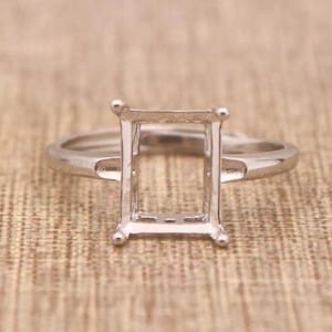 May include: A silver ring setting with a rectangular center, designed to hold a gemstone. The ring has a simple band and four prongs at each corner of the rectangular setting. The ring is shown on a textured, neutral-toned surface.